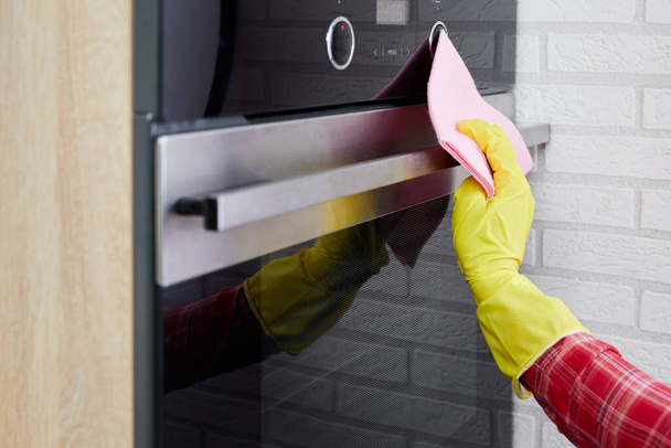 housework and housekeeping concept - close up of woman hand in protective glove with a rag cleaning oven at home kitchen - Photo, Image