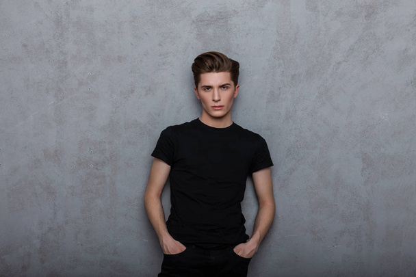 Nice modern young man hipster in a black fashionable t-shirt in jeans posing near a vintage wall indoors. Handsome guy model in stylish clothes in the studio. Youth new collection of clothes for men. - Photo, Image