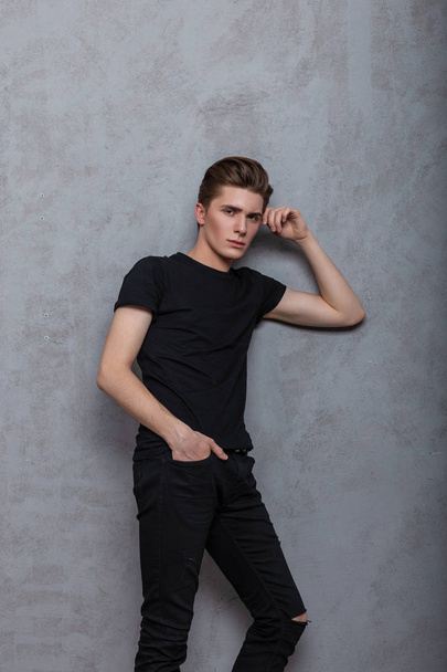 Cool handsome young man hipster in fashionable black clothes is resting near a vintage gray wall in the room. Attractive guy model with stylish hairstyle with pierced nose in trendy wear in the studio - Foto, Bild