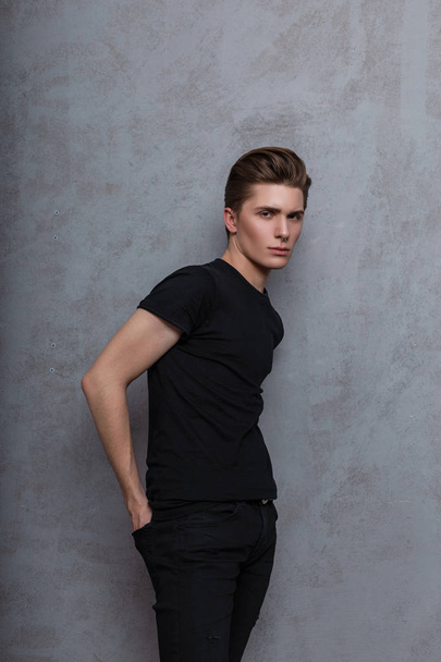Young fashionable attractive man model with a stylish hairstyle in a trendy black t-shirt in vintage jeans with a nose piercing posing near a gray wall in the studio. Modern hipster guy indoors. Style - Foto, Bild