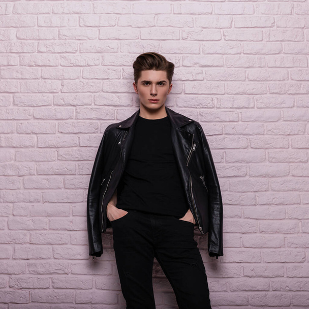 Stylish young man in black fashionable clothes with a stylish hairstyle posing indoors near a pink vintage brick wall. Handsome hipster guy in leather jacket in room. Fashion model in casual clothing. - Photo, Image