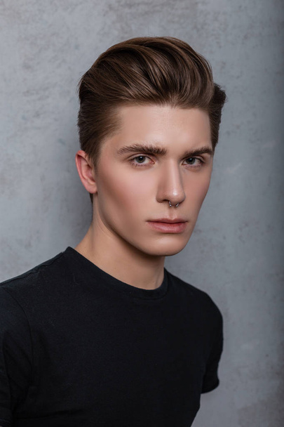 Portrait of an attractive young hipster man in a black fashionable t-shirt with a stylish hairstyle near a vintage gray wall in the studio. Close up face of handsome guy on gray background in the room - Foto, immagini