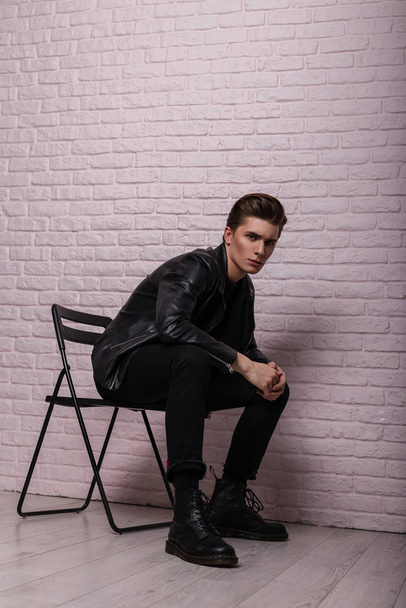 Young handsome man hipster with trendy hairstyle in fashionable black stylish youth clothes in cool leather boots is sist on a vintage chair near a pink brick wall in the studio. American trendy guy. - Photo, Image