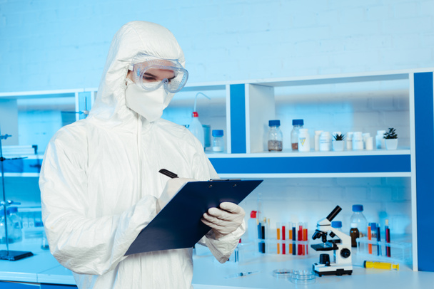 scientist in hazmat suit and goggles writing while holding clipboard near microscope  - Foto, afbeelding