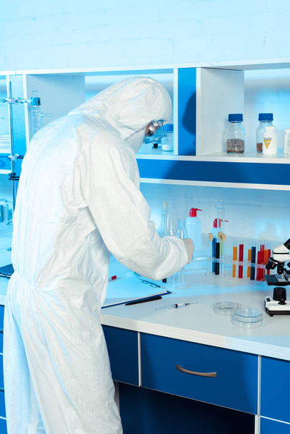 wetenschapper in hazmat pak staat in de buurt van spuit, klembord en microscoop  - Foto, afbeelding