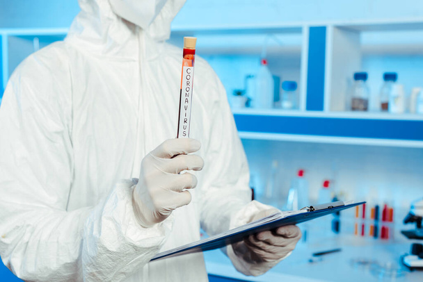 cropped view of scientist in hazmat suit holding test tube with coronavirus lettering  - Foto, Bild