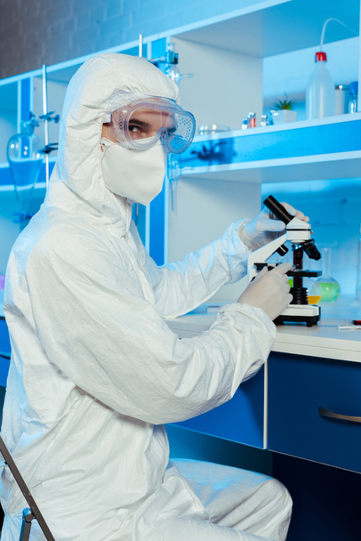 científico en traje de materiales peligrosos y gafas sentado cerca del microscopio en el laboratorio
  - Foto, Imagen