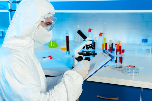 scientist in hazmat suit and goggles writing near microscope  - Fotoğraf, Görsel