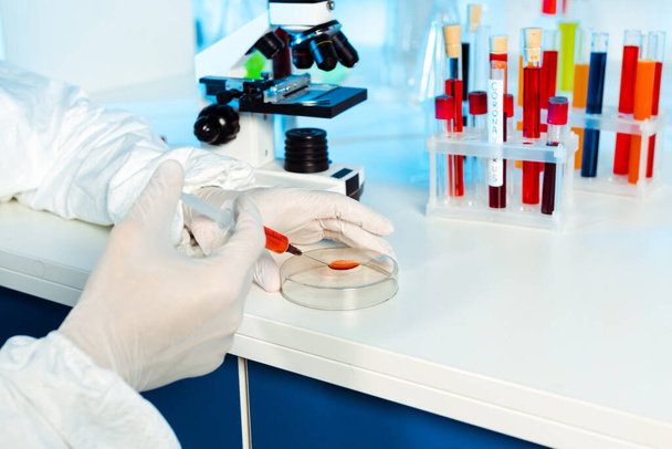 abgeschnittene Ansicht eines Wissenschaftlers in Latexhandschuhen, der eine Spritze in der Nähe einer gläsernen Testplatte hält  - Foto, Bild