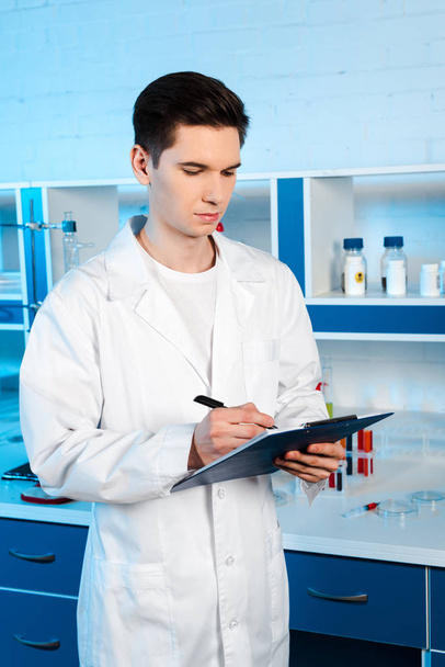 guapo científico de capa blanca sujetando portapapeles y bolígrafo en laboratorio
  - Foto, Imagen