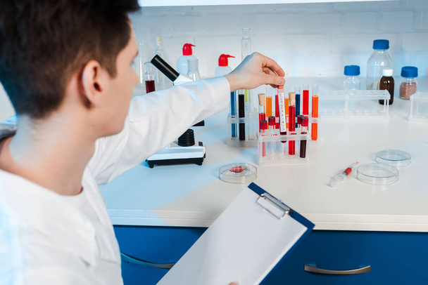 selective focus of scientist in white coat holding clipboard and touching test tube  - Φωτογραφία, εικόνα