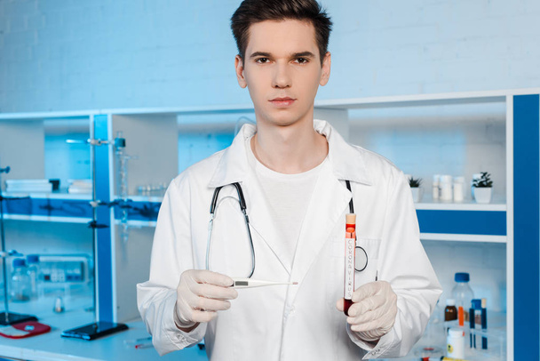 guapo científico en guantes de látex sosteniendo termómetro digital y tubo de ensayo con letras de coronavirus
  - Foto, Imagen