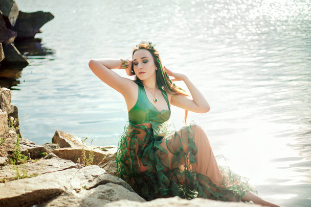Uma menina com algas no cabelo e em um vestido verde senta-se em pedras perto de uma lagoa. Modelo com uma borda com conchas. Sereia - Foto, Imagem