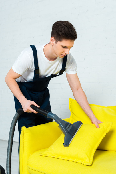 handsome cleaner in overalls removing dust on pillow with vacuum cleaner  - Fotoğraf, Görsel