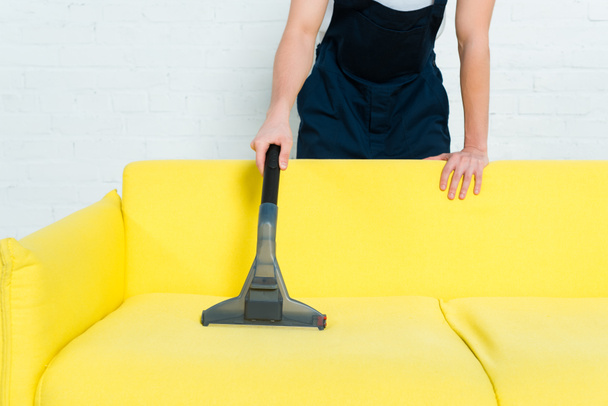 cropped view of cleaner in uniform removing dust on sofa with vacuum cleaner  - Foto, Imagen