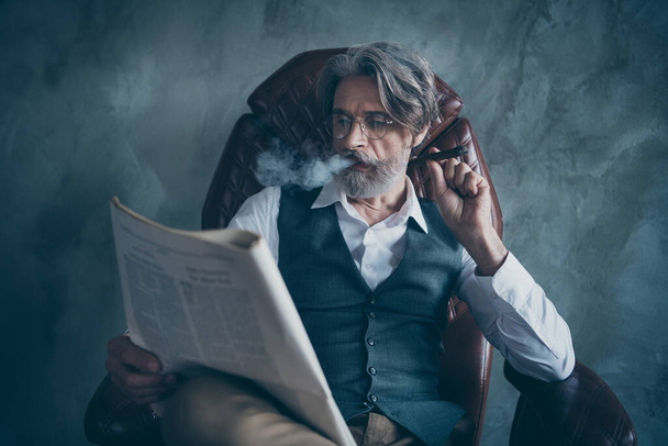 World economy political news concept.Luxury classy chic dandy old man company owner morning newspaper reader sit chair smoke cigar wear shirt brown pants trousers isolated grey color background - 写真・画像
