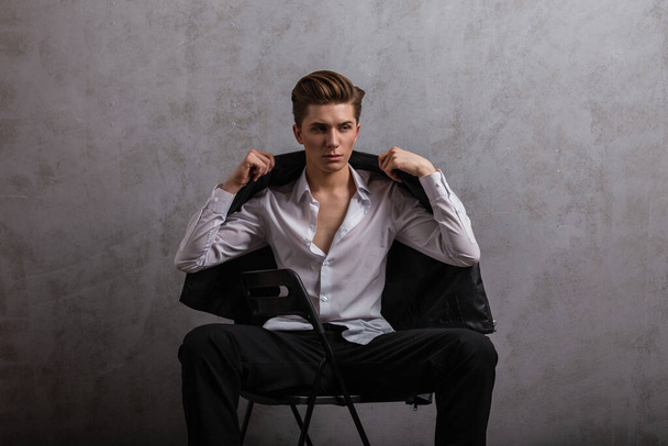 Cool handsome young man hipster puts on a fashionable leather black jacket. Attractive guy in stylish clothes posing on a chair near a vintage gray wall indoors. American style. Fashion wear for men. - Foto, immagini