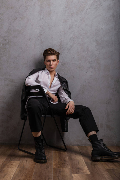 Modieus model van een Amerikaanse jongeman in een zwart leren jasje in een klassiek wit shirt in trendy schoenen zit op een stoel in de buurt van een vintage grijze muur in de kamer. Elegante jongen in jeugdkleding binnen - Foto, afbeelding