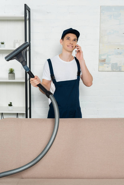 alegre limpiador en gorra y overoles sosteniendo la aspiradora y hablando en el teléfono inteligente
  - Foto, imagen