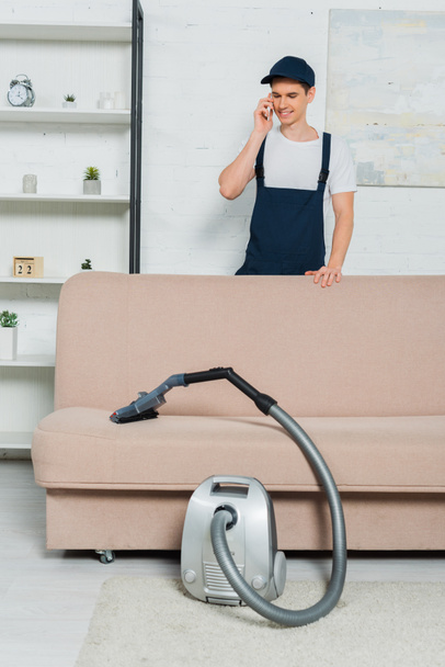 happy young cleaner in cap and overalls holding vacuum cleaner and talking on smartphone  - Foto, immagini