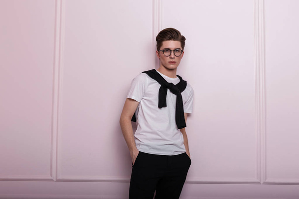 Model of a elegant young man in a glasses in a stylish white t-shirt in fashionable jeans with a piercing with a trendy hairstyle stands near a pink wall in the room. Handsome hipster guy indoors. - Photo, Image