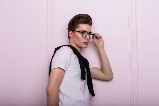 Όμορφος Αμερικανός νεαρός hipster σε λευκά κομψά ρούχα με μοντέρνο χτένισμα σε γυαλιά που ποζάρουν σε ένα δωμάτιο κοντά σε ένα ροζ τοίχο. Πολύ ελκυστικό αντρικό μοντέλο μόδας σε casual ντύσιμο στο στούντιο. - Φωτογραφία, εικόνα