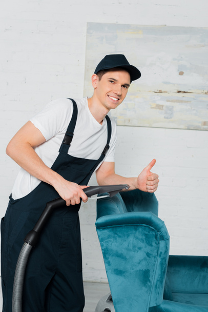 happy cleaner in cap showing thumb up while dry cleaning blue armchair with vacuum cleaner  - Photo, Image