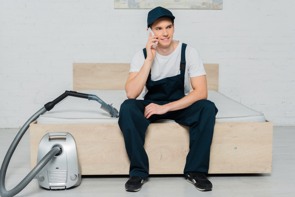 happy cleaner in cap sitting on bed and talking on smartphone near vacuum cleaner  - Foto, imagen