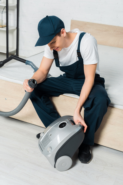 happy cleaner in cap holding hose near vacuum cleaner in bedroom  - Foto, Imagen