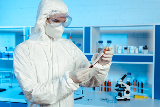 scientist in hazmat suit, latex gloves and goggles holding test tube with coronavirus lettering  - Valokuva, kuva