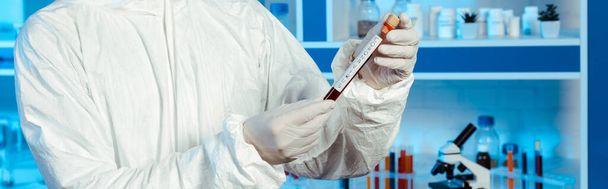 plano panorámico de científico en traje de materiales peligrosos y guantes de látex sosteniendo tubo de ensayo con letras de coronavirus
  - Foto, Imagen