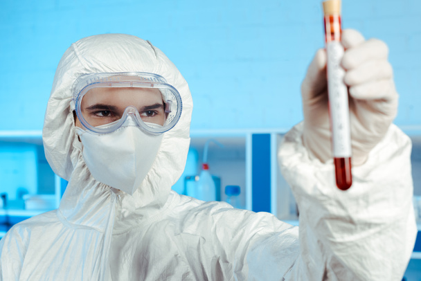 selective focus of scientist in hazmat suit and goggles holding test tube with coronavirus lettering  - Foto, afbeelding