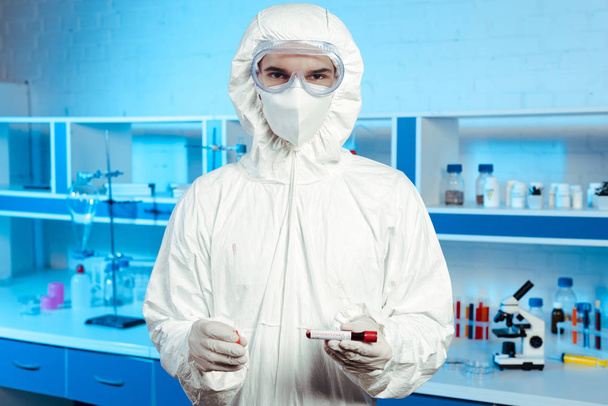 vegyvédelmi ruhában, orvosi maszkban és védőszemüvegben lévő tudós, aki a mintát koronavírus felirattal tartja  - Fotó, kép