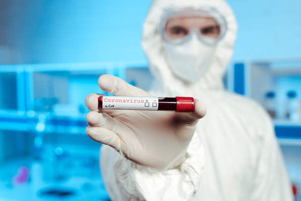 selective focus of scientist in hazmat suit, medical mask and goggles holding sample with coronavirus lettering  - Foto, afbeelding