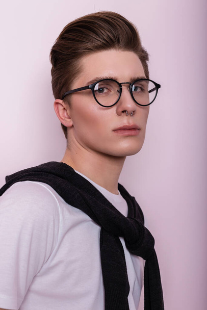 Zbliżenie studio portret młodego modnego hipstera z czystą skórą z kolczykami w vintage biały t-shirt na różowym tle. sexy przystojny facet hipster w a okulary z fryzura wewnątrz - Zdjęcie, obraz
