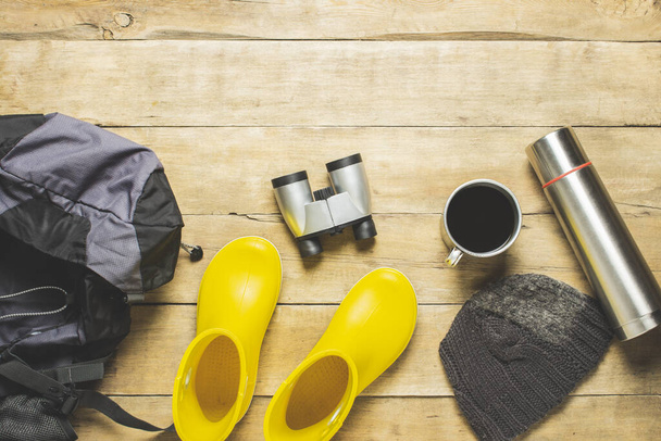 Yellow rain boots, backpack, binoculars, camping equipment on a wooden background. The concept of hiking, tourism, camp, mountains, forest. Banner. Flat lay, top view. - Photo, image