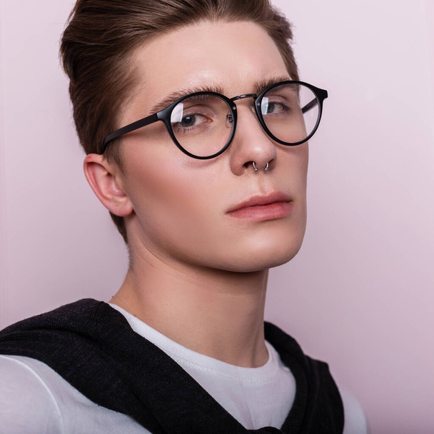 Portrait of an attractive young hipster man in a glasses in a white fashionable t-shirt with a hairstyle near a vintage wall. Close up face of american guy on pink background - Фото, зображення