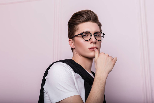 Sexy European model of a young man in fashionable glasses with a stylish hairstyle with piercings in a white T-shirt posing near a pink vintage wall indoors. Studio portrait of a handsome hipster guy. - 写真・画像