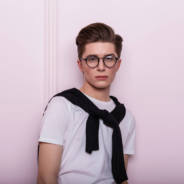 Portrait élégant d'un beau jeune homme hipster avec une peau propre et saine dans des lunettes à la mode avec un nez percé dans un T-shirt blanc à la mode avec un cardigan près d'un mur vintage rose. Élégant gars à l'intérieur
. - Photo, image