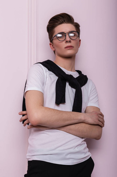 Μοντέρνος κομψός νεαρός hipster άντρας με χτένισμα με μοντέρνα γυαλιά σε λευκό vintage t-shirt με μαύρη ζακέτα να ποζάρει κοντά σε έναν μοντέρνο ροζ τοίχο εσωτερικά. - Φωτογραφία, εικόνα