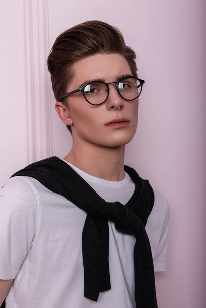 Retrato de estúdio elegante de um jovem hipster bonito com pele limpa e saudável em óculos da moda com piercing no nariz em uma camiseta branca com um casaco de lã em um fundo de parede vintage rosa. Rapaz americano
. - Foto, Imagem