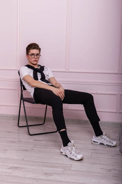 Moderno bonito modelo jovem na moda em um óculos elegantes em uma camiseta branca na moda em jeans vintage em um tênis se senta em uma cadeira perto de uma parede rosa no estúdio. Inglês hipster cara dentro de casa
. - Foto, Imagem