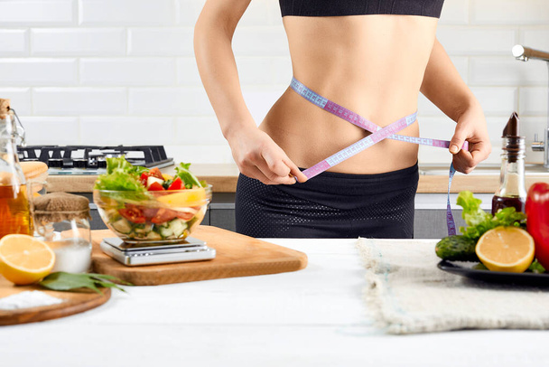 Diet, healthy eating, food and weigh loss concept. Young woman measuring waist near tomatoes, peppers and salad on the kitchen interior. - Photo, image