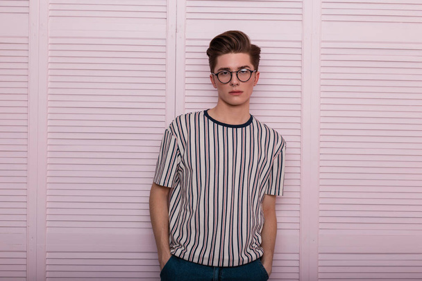 Moderni seksikäs nuori mies hipster muodikas lasit kampauksen trendikäs raidallinen t-paita seistä lähellä vintage puinen vaaleanpunainen seinä studiossa. Tyylikäs komea moderni mies. Englanti muoti
. - Valokuva, kuva