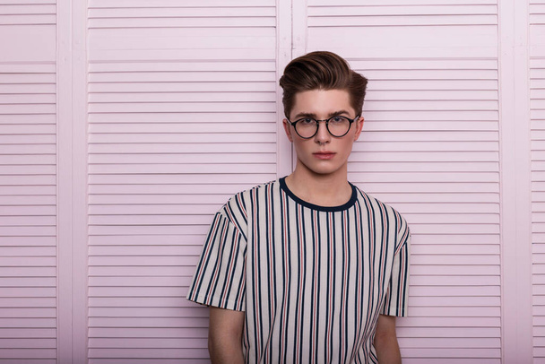 Moderne élégant jeune homme hipster dans des lunettes à la mode avec une coiffure dans un t-shirt à rayures à la mode posant près d'un mur vintage en bois rose dans le studio. Portrait d'un homme séduisant. Mode anglaise
 - Photo, image