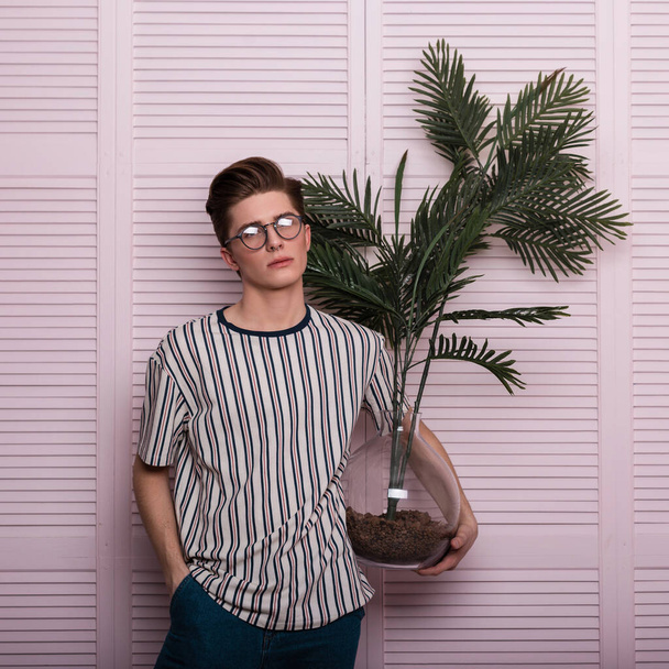 Modischer junger Hipster in stylischem T-Shirt mit Vintage-Brille und einer exotischen Kunstblume im Glastopf steht auf einem Stuhl neben einer Holzwand im Atelier. Attraktives Modell in Innenräumen. - Foto, Bild