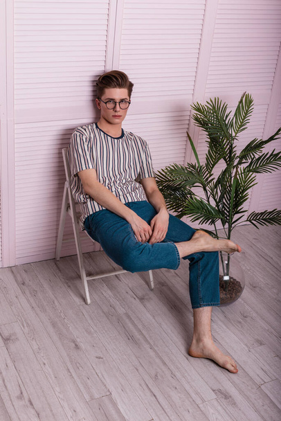 European young man with a fashionable hairstyle in stylish youth clothes in vintage glasses is resting on a chair next to a green plant near a wooden pink wall in the room. Trendy guy fashion model. - Fotoğraf, Görsel