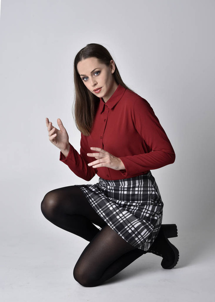 full length portrait of a pretty brunette girl wearing a red shirt and plaid skirt with leggings and boots. Seated pose on a  studio background. - Photo, Image