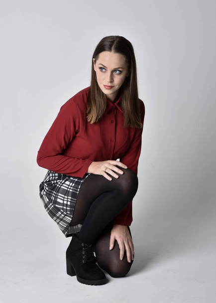 full length portret van een mooi brunette meisje met een rood shirt en geruite rok met legging en laarzen. Zittende houding op een studio achtergrond. - Foto, afbeelding