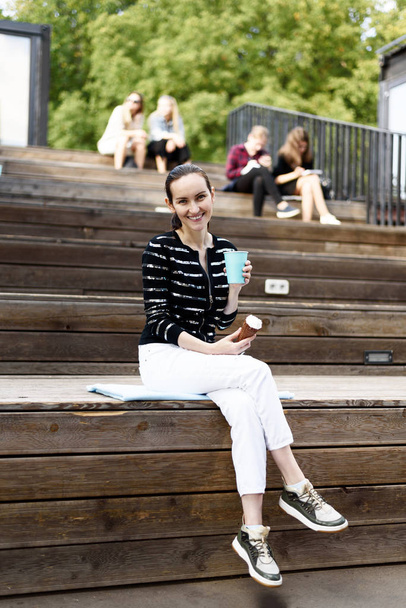Dunkelhaariges Mädchen mit Eis und blauem Glas Kaffee in der Hand sitzt auf Bank im Sommerpark, Freizeitkonzept - Foto, Bild